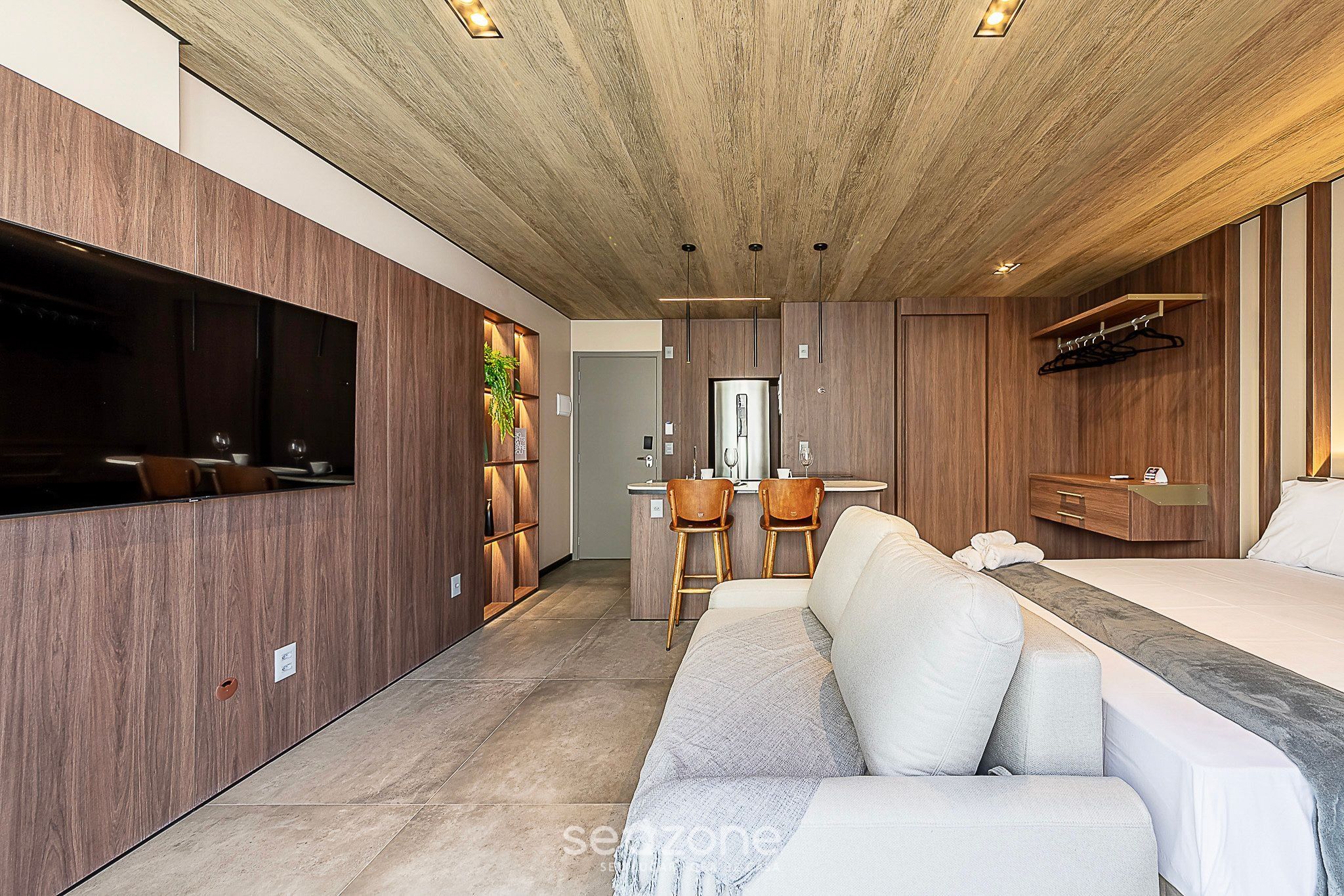Una habitación de hotel moderna con techo de madera, un televisor de pantalla plana grande, un sofá, una cama, una pequeña zona de cocina y un armario de madera.