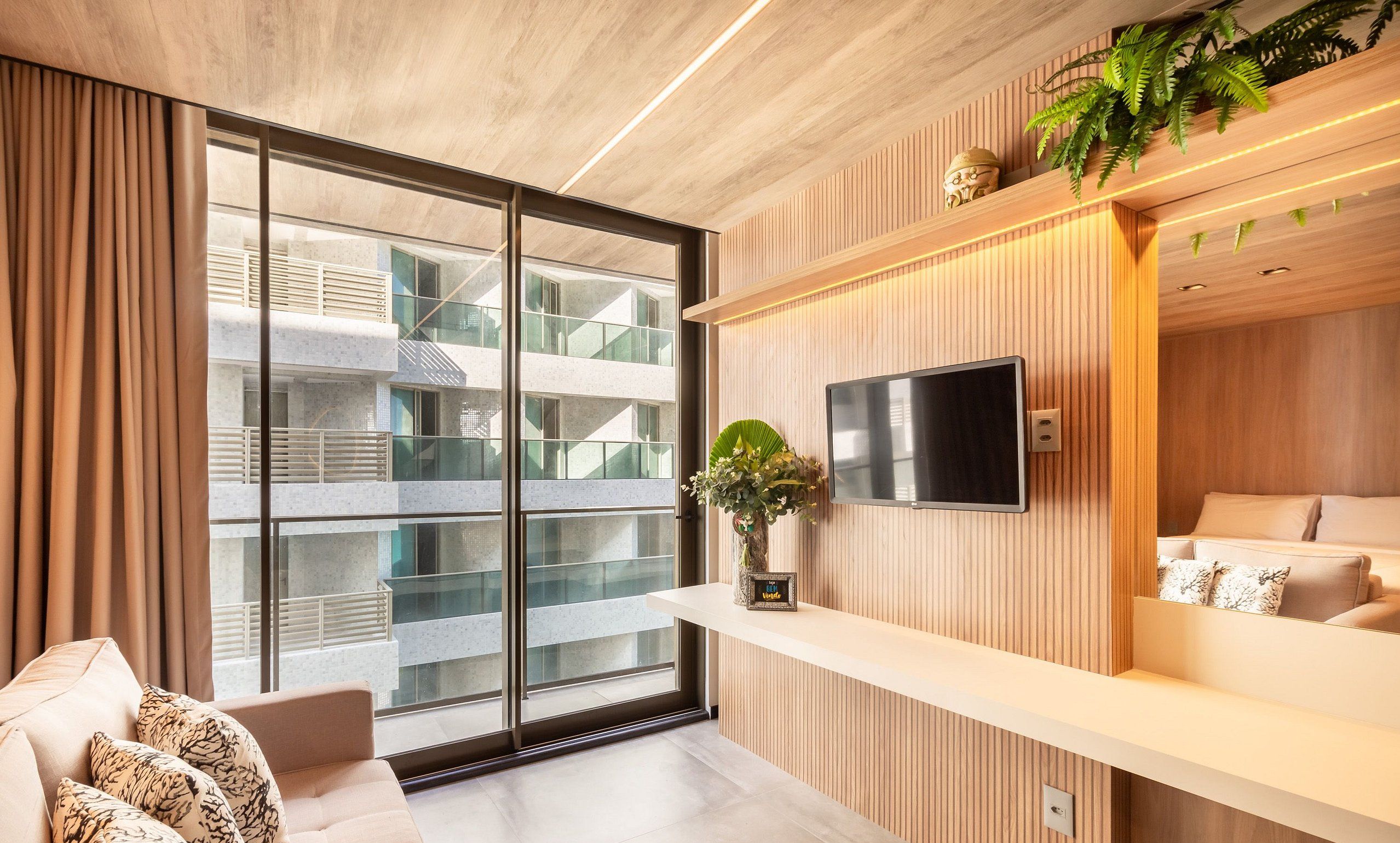 A modern hotel room with a large sliding glass door, a flat-screen TV, a sofa, and a small plant on a shelf.
