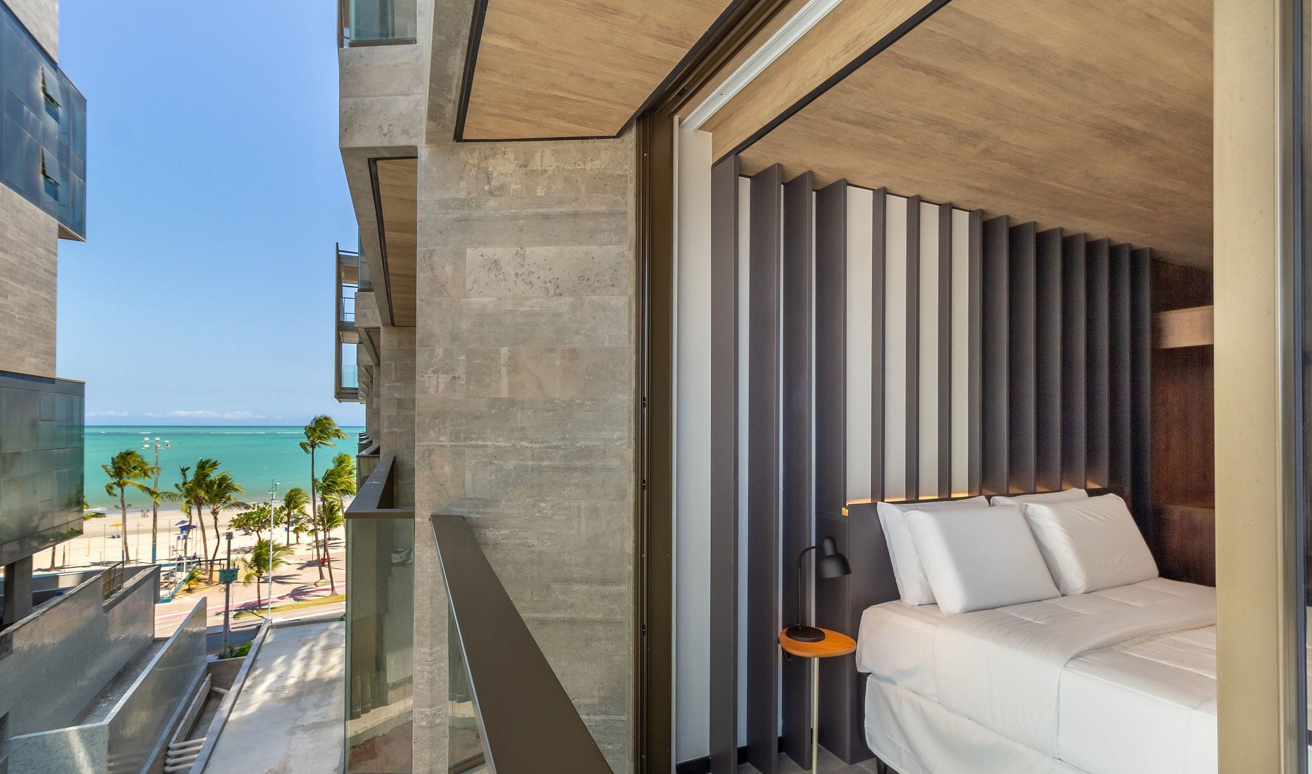 A hotel room with a bed, a nightstand, and a view of the beach and ocean.