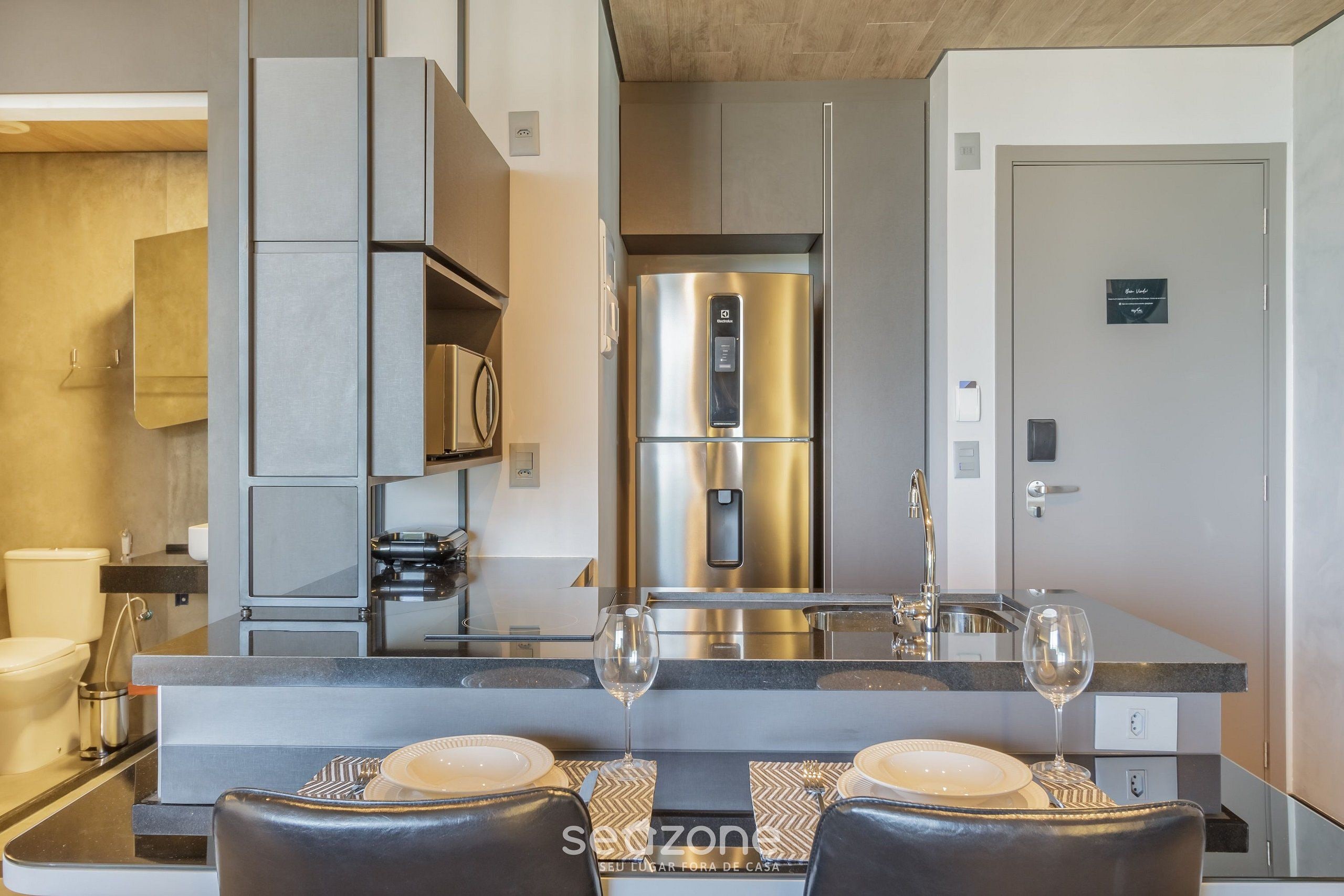 Une cuisine moderne équipée d’un réfrigérateur en acier inoxydable, d’un four encastré et d’un évier. On y trouve également un coin repas avec une table dressée pour deux personnes, deux verres à vin et une chaise.