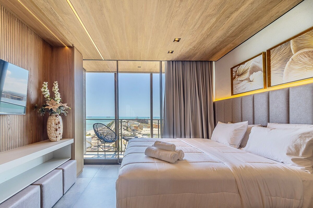 Ein modernes Hotelzimmer mit einem großen Bett, einem Fernseher, einem Balkon mit Blick auf den Strand und einer dekorativen Vase.