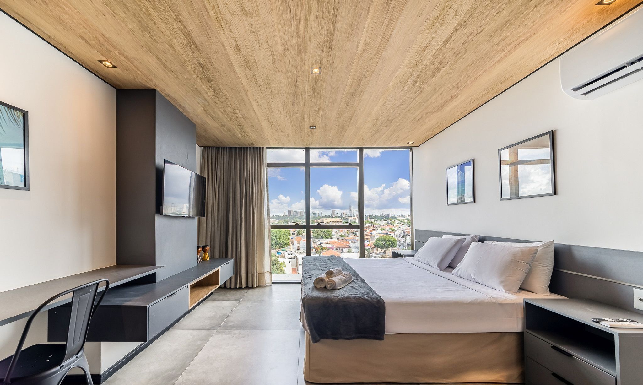Un dormitorio moderno con una cama grande, un escritorio y un televisor. Se ve un paisaje urbano a través de ventanas amplias.