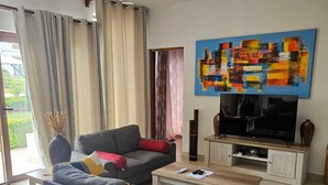 Un salon moderne avec un canapé gris, une table basse et un téléviseur à écran plat posé sur un meuble en bois. On y trouve également un ventilateur de plafond, un grand tableau abstrait et une vue sur l’extérieur grâce à une porte vitrée.
