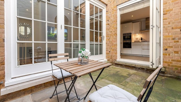 Un patio con una mesa y sillas de madera, un banco y vista a una cocina a través de un ventanal grande.