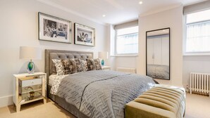 Un dormitorio moderno con una cama grande, una mesita de noche con lámpara, un banco y dos cuadros en la pared.