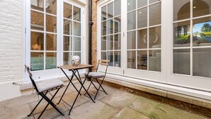 Un patio con una mesa y dos sillas, amplias ventanas con vistas a la vegetación y un muro de ladrillo.