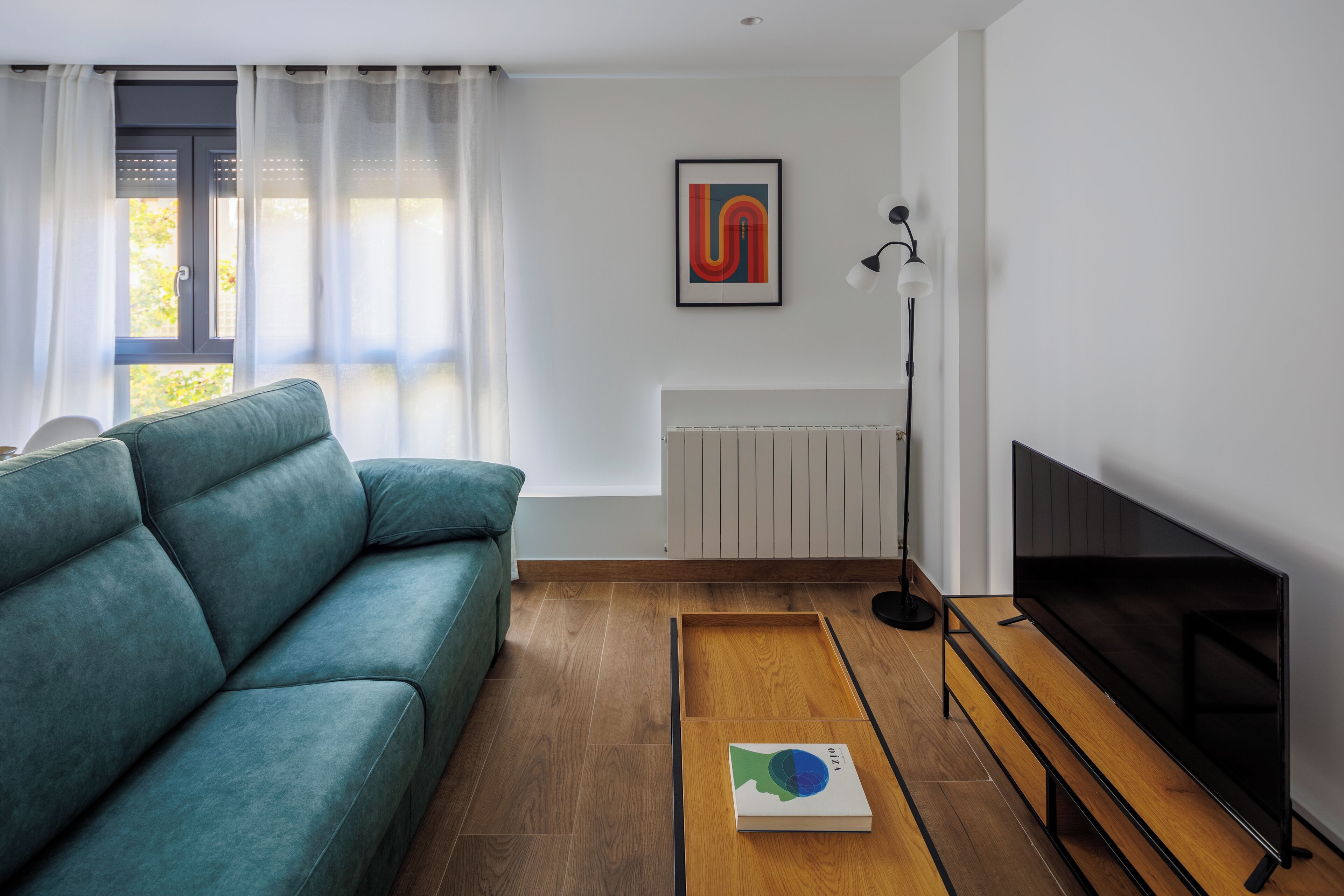 Ein modernes Wohnzimmer mit einem petrolfarbenen Sofa, einem Couchtisch aus Holz mit einem Buch, einem Flachbildfernseher und einer Heizung.