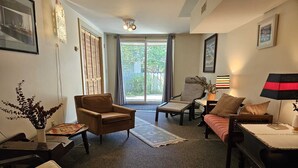 Una sala de estar con un sillón marrón, un sofá, una mesa de centro y una lámpara. Se ve una ventana con cortinas, una puerta y una planta a la izquierda.