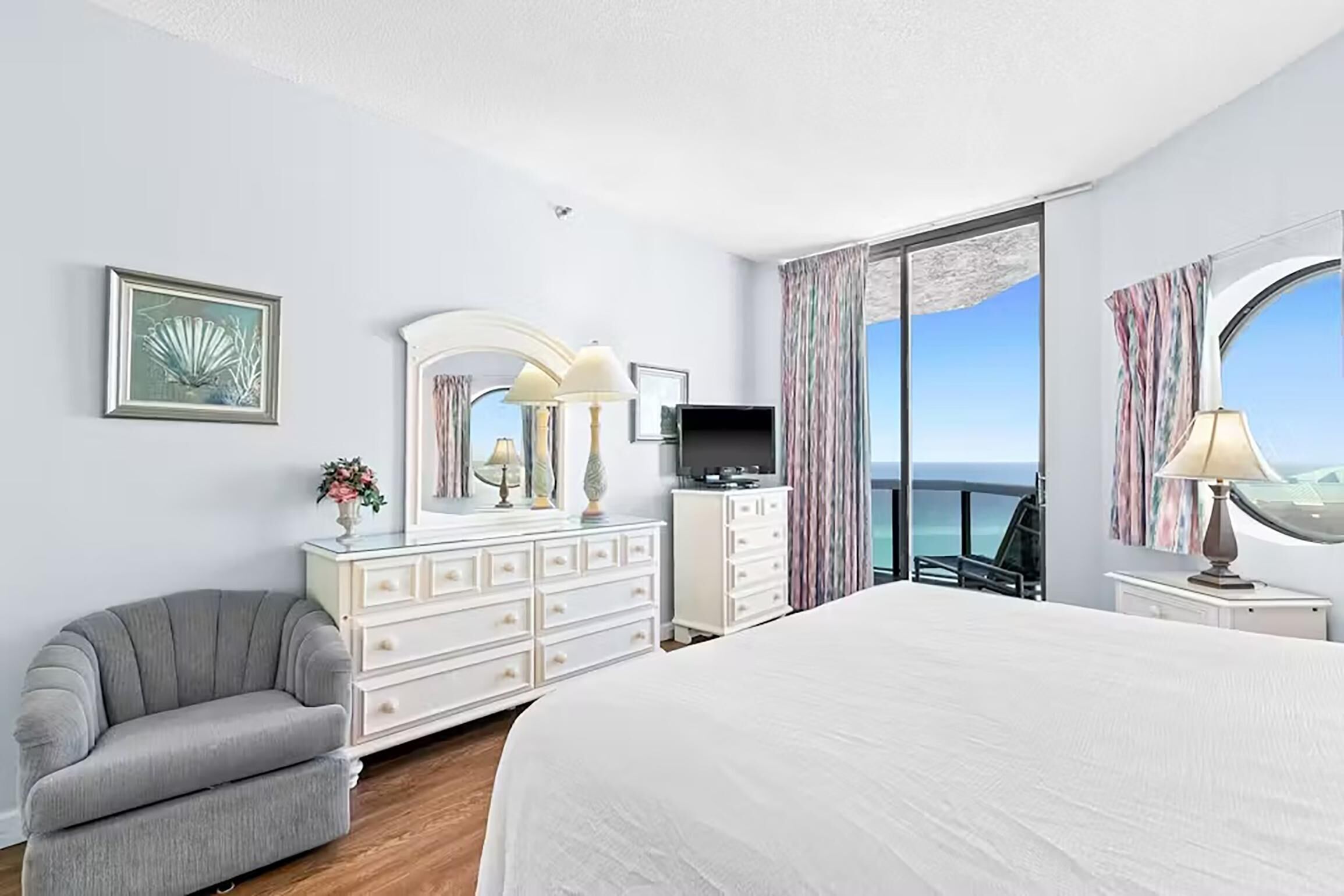A hotel room with a large bed, a dresser, a vanity with a mirror, a television, and a view of the ocean through a large window.
