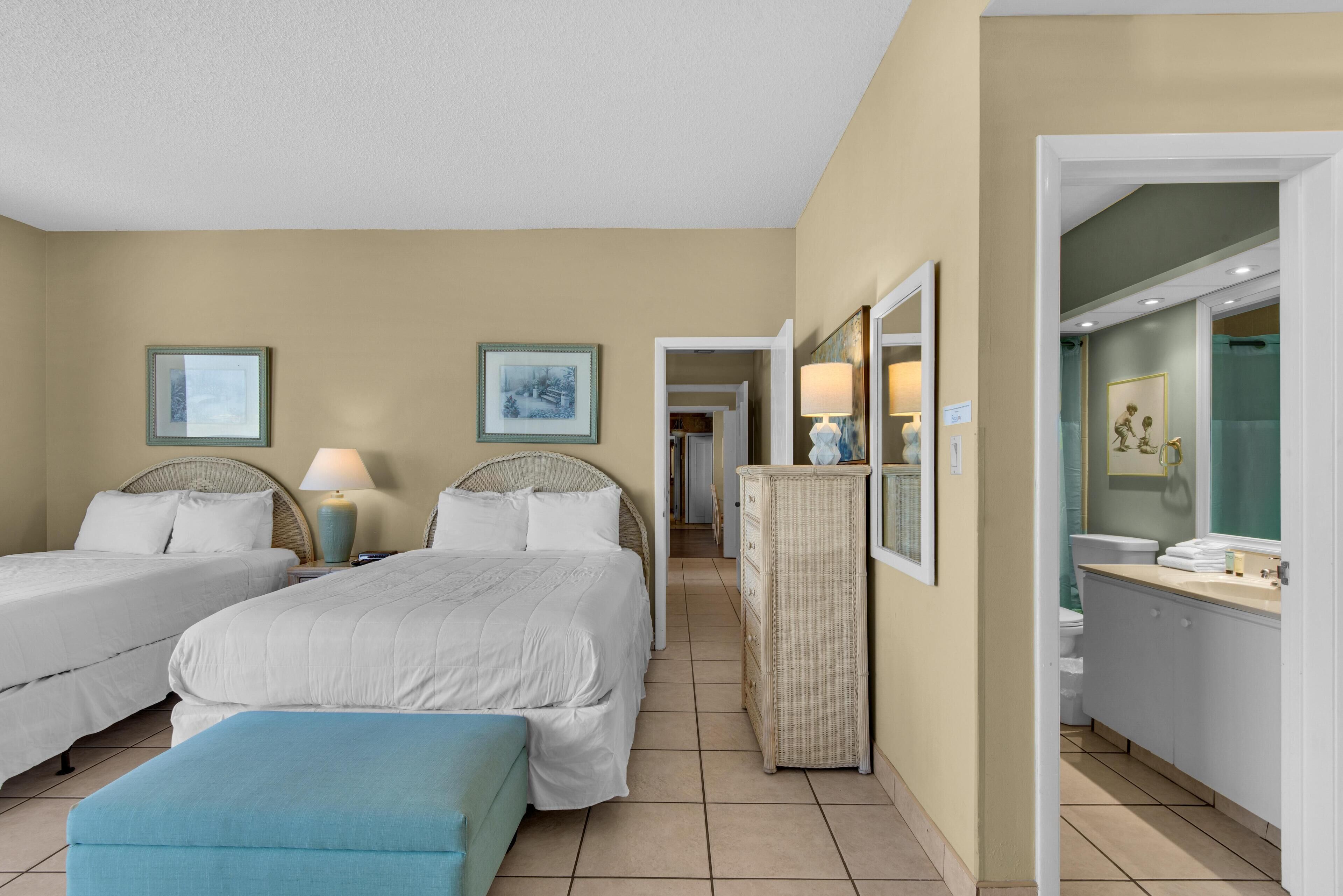A hotel room with two beds, a sitting area, a bathroom with a vanity, and a hallway leading to other rooms.