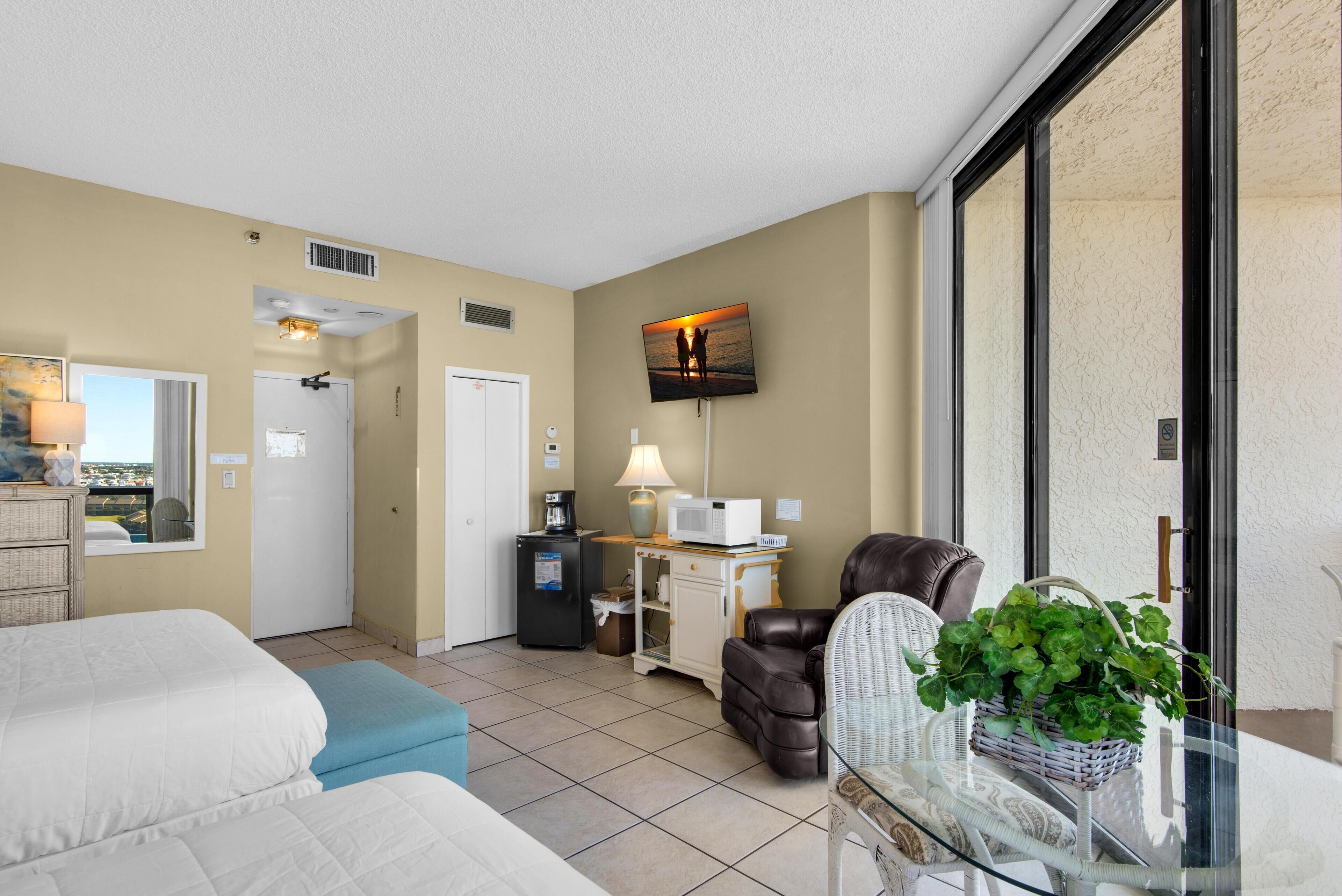 A hotel room with a bed, a massage chair, a desk with a lamp, a TV on the wall, and a view of the outside.