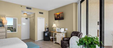 A hotel room with a bed, a massage chair, a desk with a lamp, a TV on the wall, and a view of the outside.