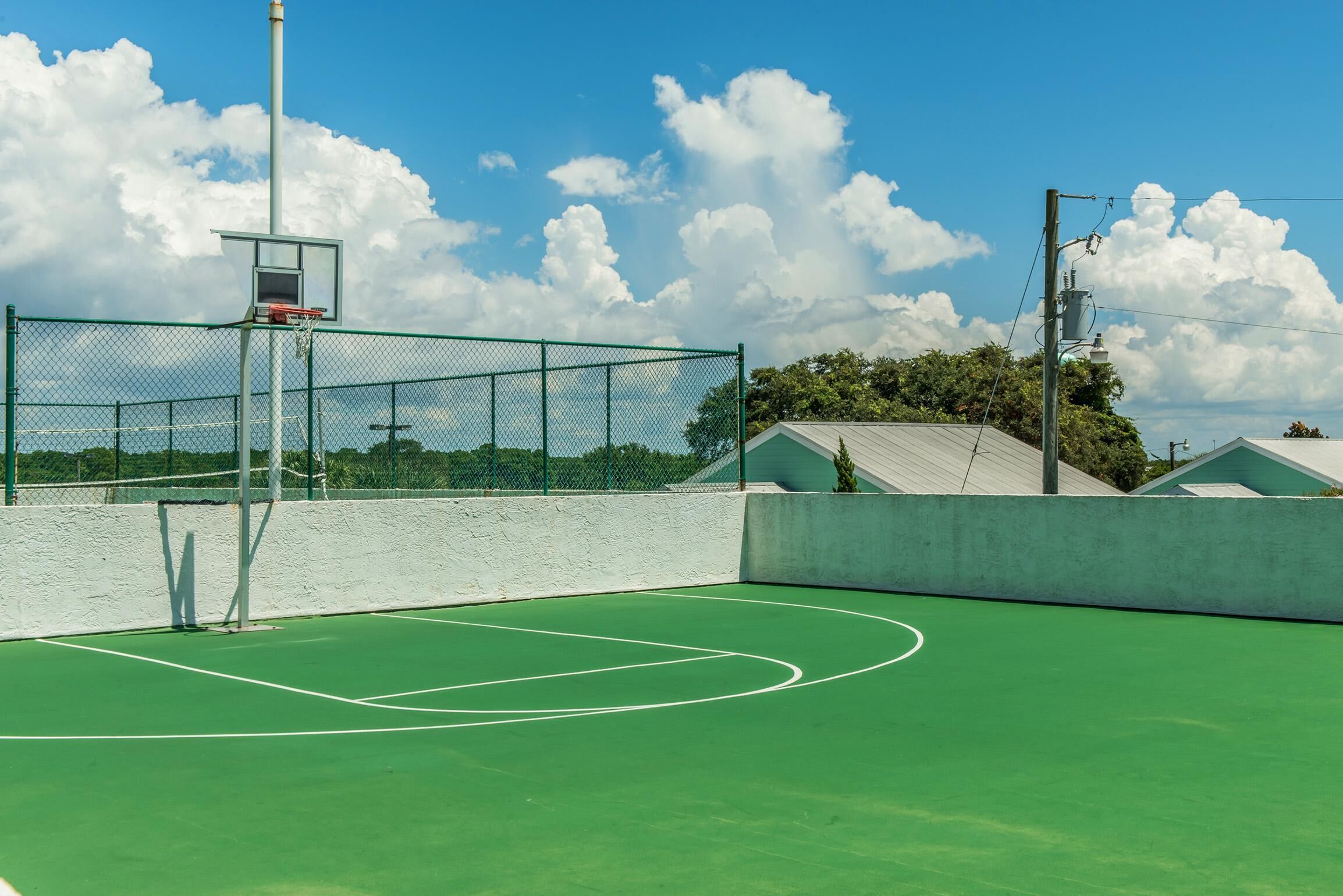 Ein Basketballplatz im Freien mit Korb, umgeben von einem grünen Zaun und einer weißen Mauer.