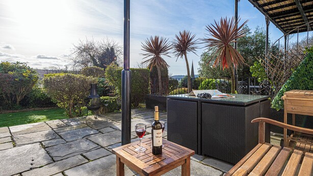 Een terras met een stenen vloer, tuinmeubilair en een fles wijn op een tafel.