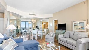 A spacious living room with a blue sofa, a grey sofa, and a glass coffee table. There is a dining area in the background with a chandelier. - Surfside Resort #1508 (Miramar Beach)
