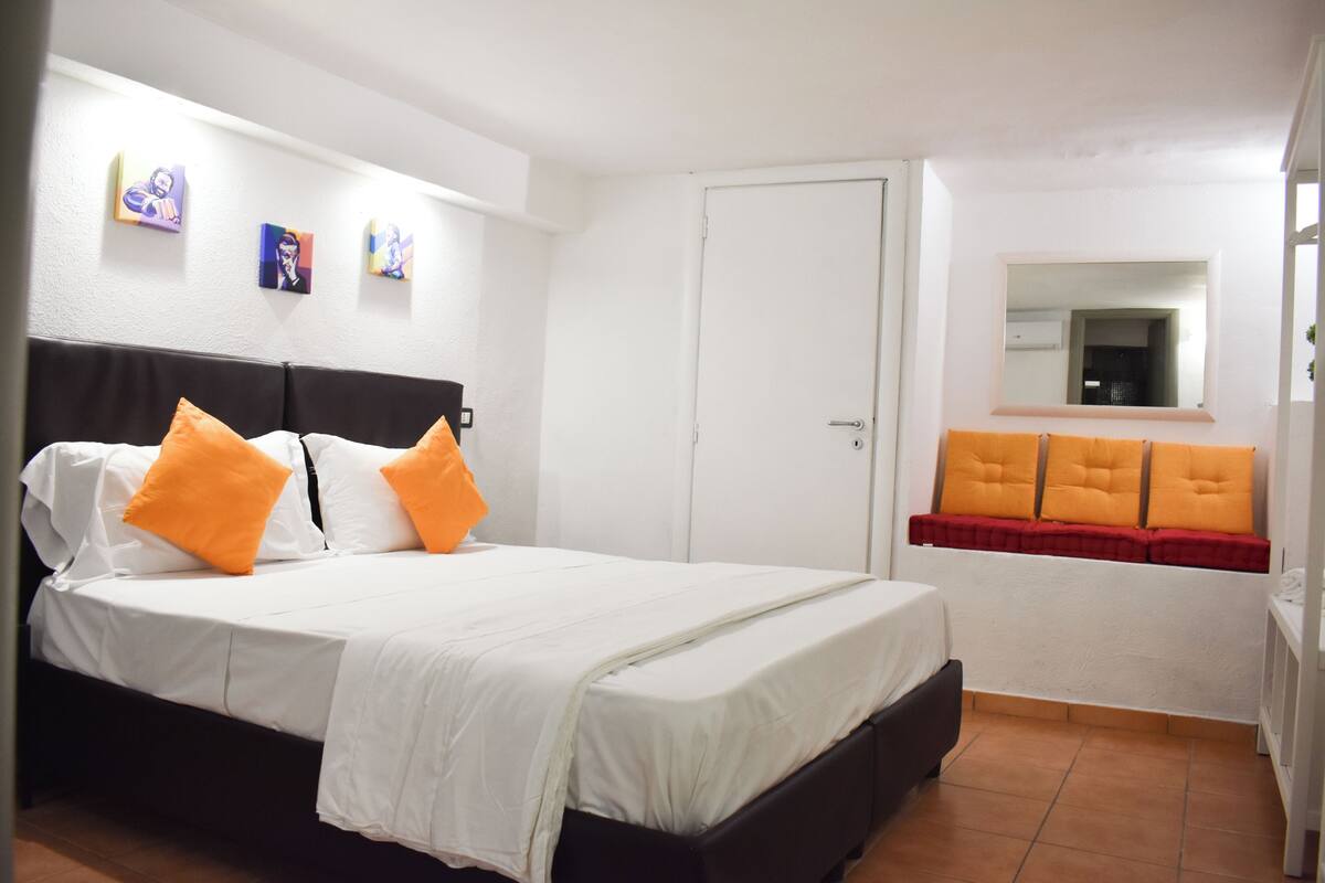 A neatly arranged bedroom with a bed, a wardrobe, and a small seating area with cushions.