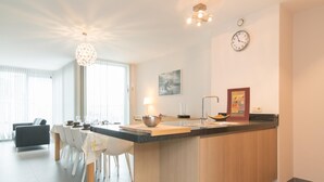A modern kitchen with a central island, a dining area with white chairs, and a living room with a dark sofa. - Penthouse in Ostend With sea View Dining (Ostend)