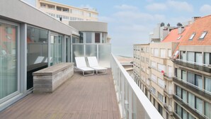A balcony with two white lounge chairs, a wooden deck, and a view of the sea and buildings. - Penthouse in Ostend With sea View Dining (Ostend)