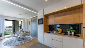 A modern kitchen with white cabinets, a wooden countertop, and a built-in oven. There is a dining area with a round table and chairs, and a view of the city through large windows. - Apartment Leonarda (Zadar)