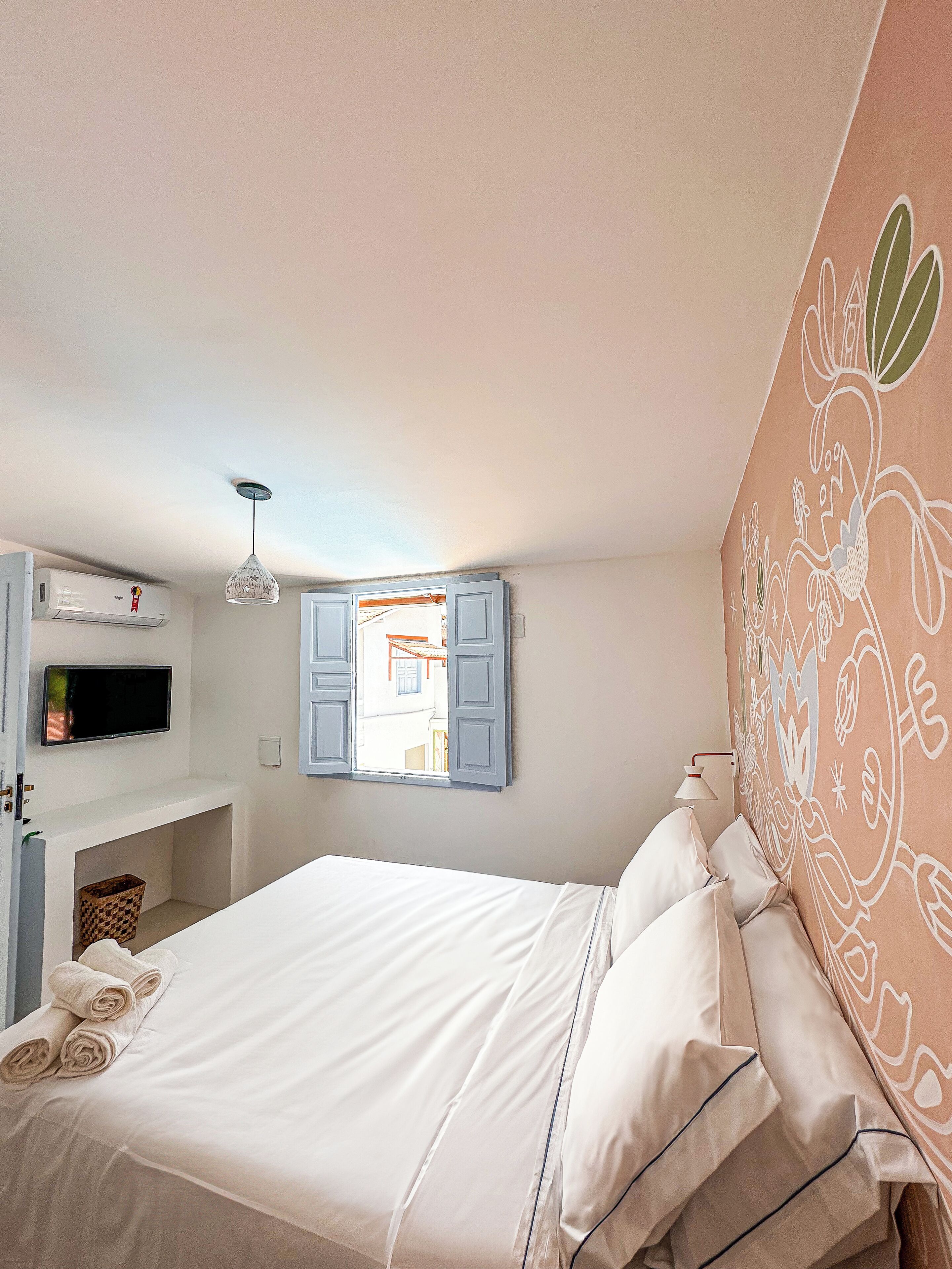 A bedroom with a bed, a TV, an air conditioning unit, a window with open shutters, and a wall with a floral pattern.