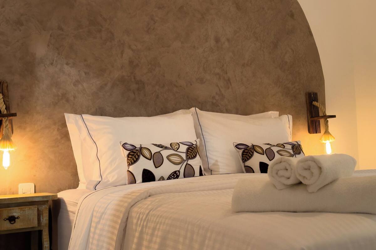 Une chambre à coucher avec un grand plafond en arc de cercle, du linge de lit blanc, des coussins à motifs et des lampes de chevet.