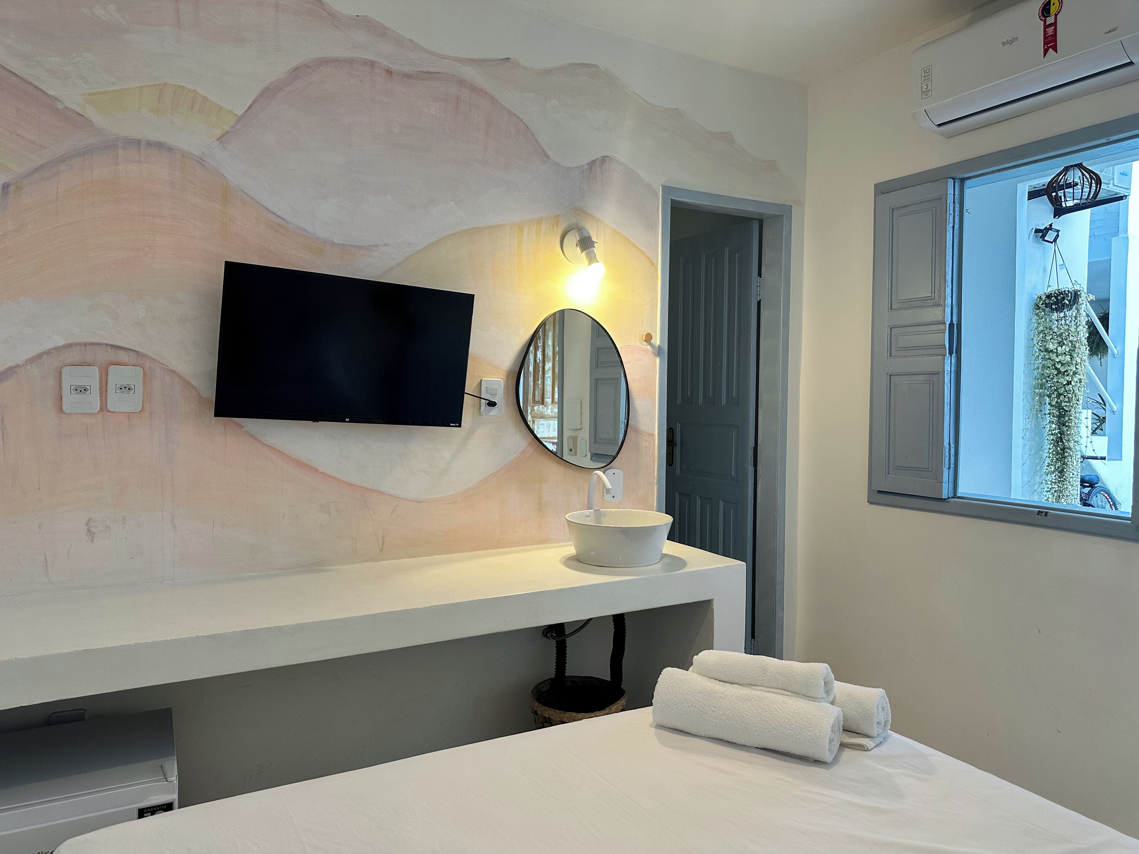 A modern bathroom with a mounted TV, a round mirror, a sink, and a window with a view of greenery.