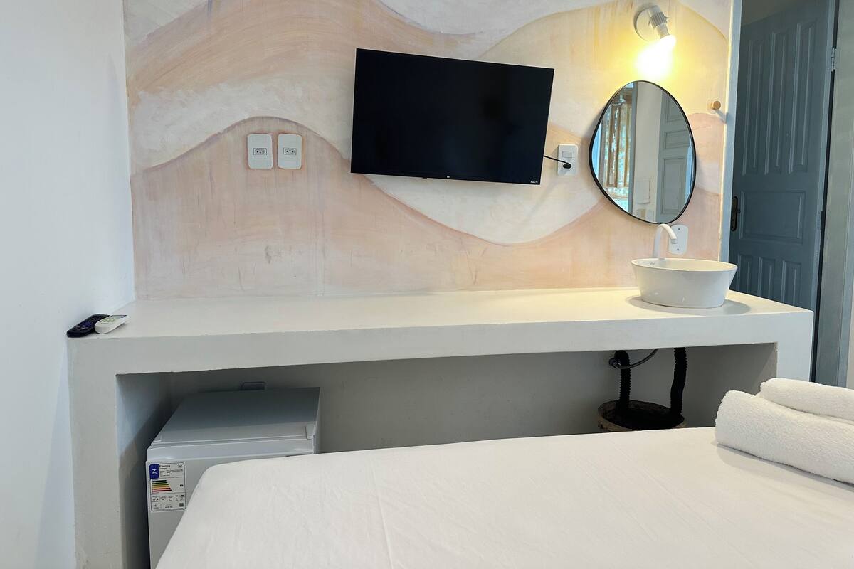 Une chambre d’hôtel avec un lit, une table de chevet blanche ornée d’un miroir rond, un téléviseur à écran plat fixé au mur et un lavabo blanc.