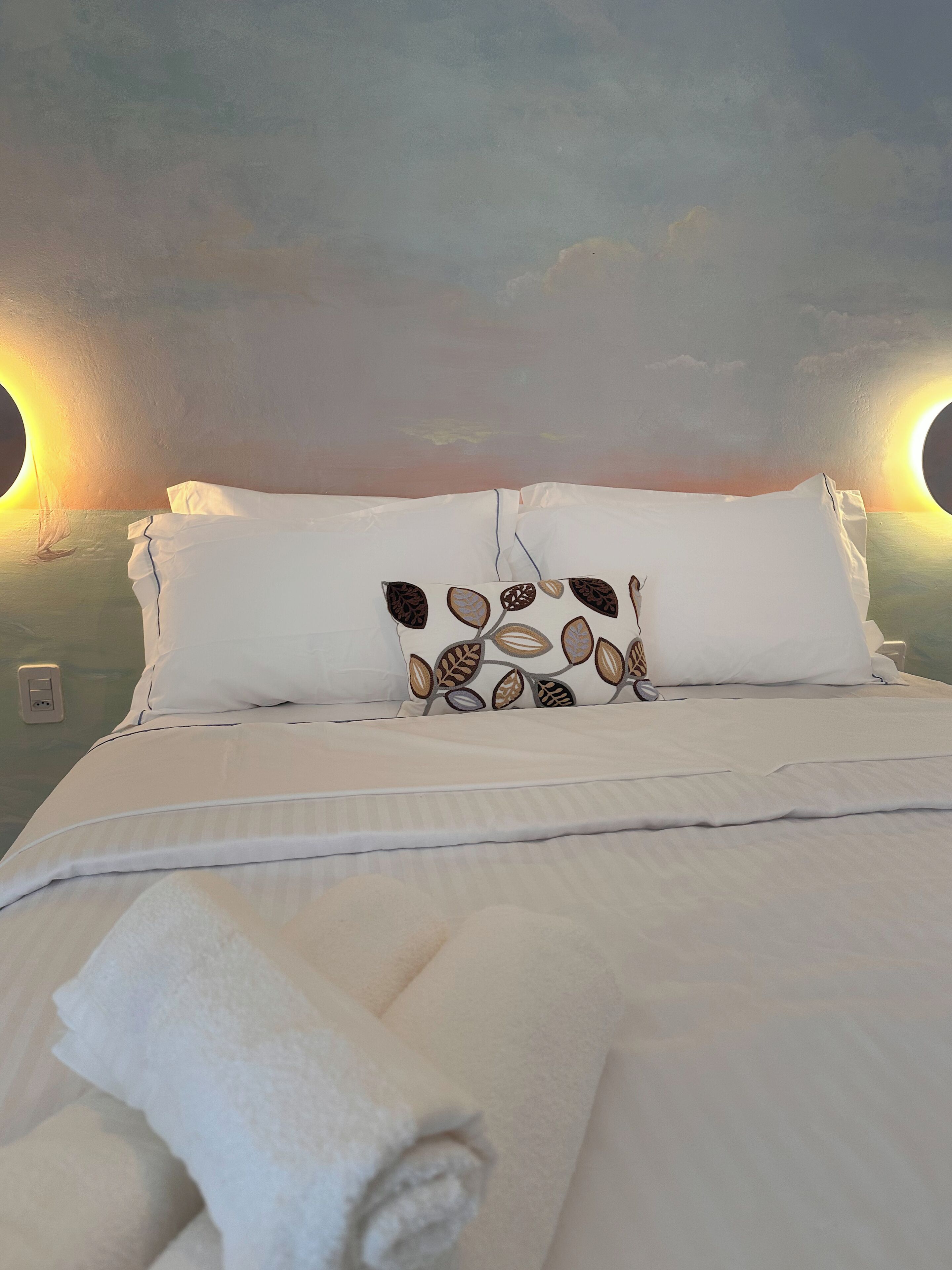 Una cama bien tendida con sábanas blancas y un cojín decorativo con motivos de hojas. Hay dos lámparas de pared a cada lado de la cama.