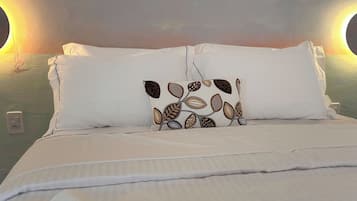 Una cama bien tendida con sábanas blancas y un cojín decorativo con motivos de hojas. Hay dos lámparas de pared a cada lado de la cama.