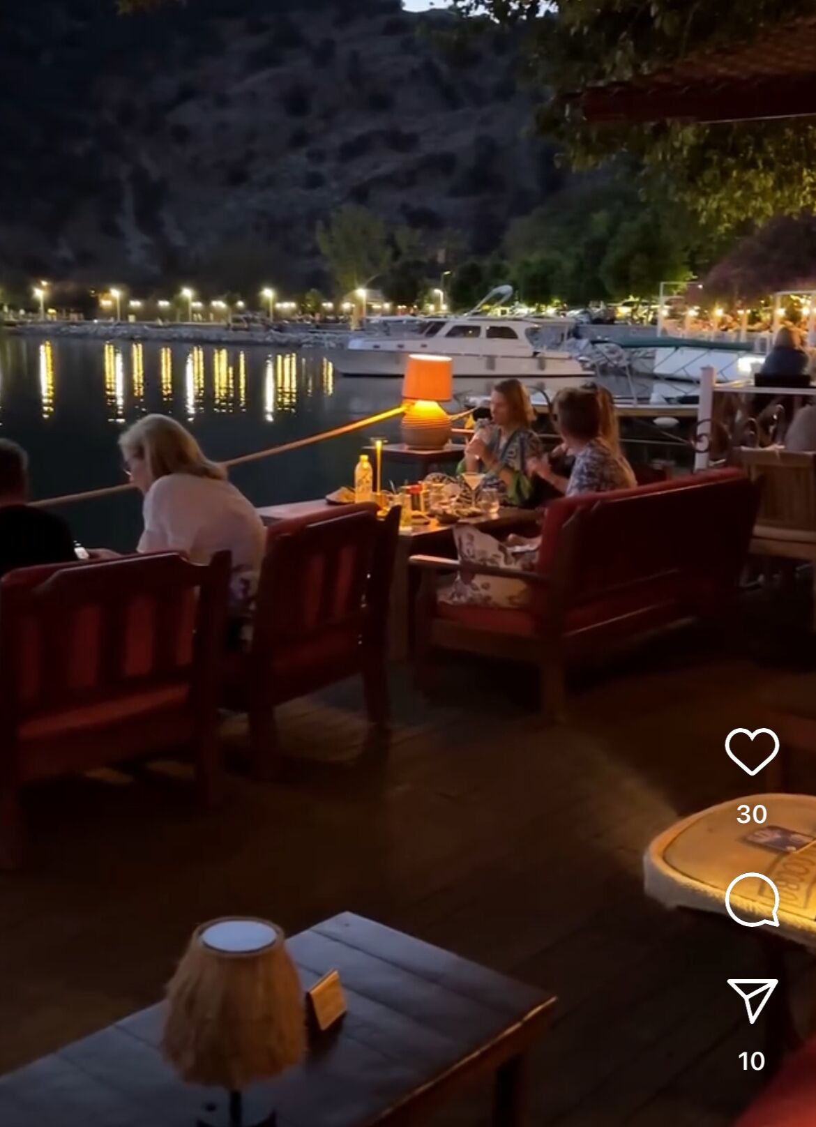 An outdoor dining area with wooden furniture and a table lamp, overlooking a marina with boats and a calm body of water.