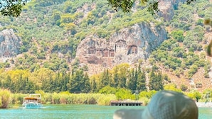 A person seated at a table with a drink, overlooking a lake with a boat and ancient ruins in the background. - Vome (Ortaca)