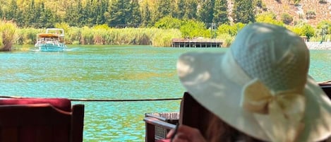 Una persona sentada a una mesa con una bebida, con vista a un lago que incluye una embarcación y ruinas antiguas al fondo.