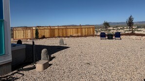 A patio with a wooden fence, two blue chairs, and a gravel ground. - Charming Desert Beach House Tiny home, with AC, WiFi, game room/fitness room. (Beryl)