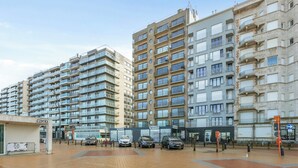 Een meerlaags woongebouw met balkons, een entree op de begane grond en een parkeerplaats met auto's.