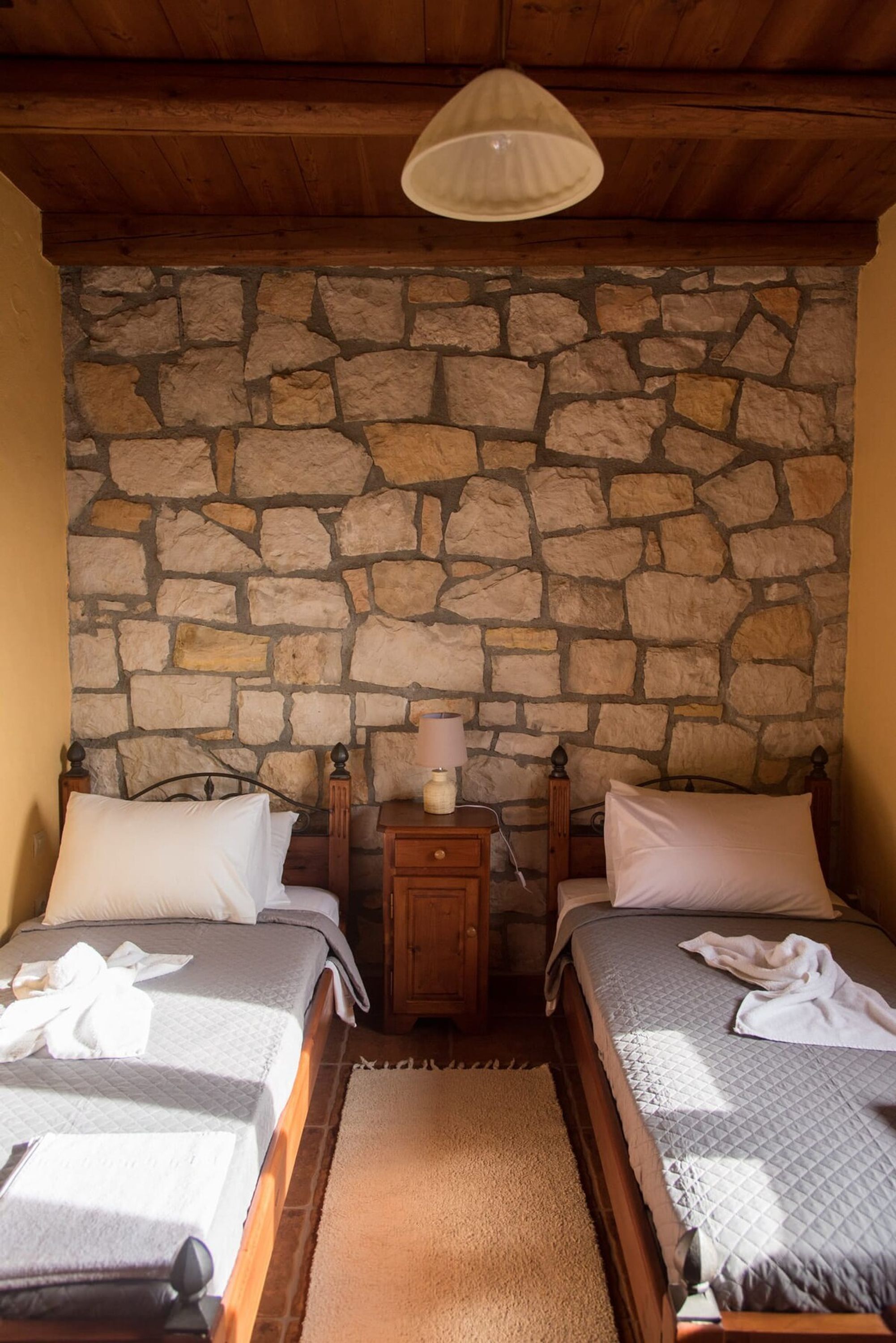 A room with two beds, a stone wall, a wooden ceiling, and a nightstand with a lamp.