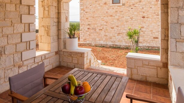 Un patio con paredes de piedra, una mesa y sillas de madera, un cuenco de fruta y plantas en macetas.
