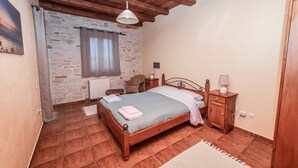 Un dormitorio con pared de piedra, vigas de madera en el techo, una cama con colcha azul, una mesita de noche con lámpara y una ventana con cortinas.