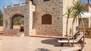 Un edificio de piedra con techo de tejas, un arco y una zona de piscina con sillones de descanso y palmeras.