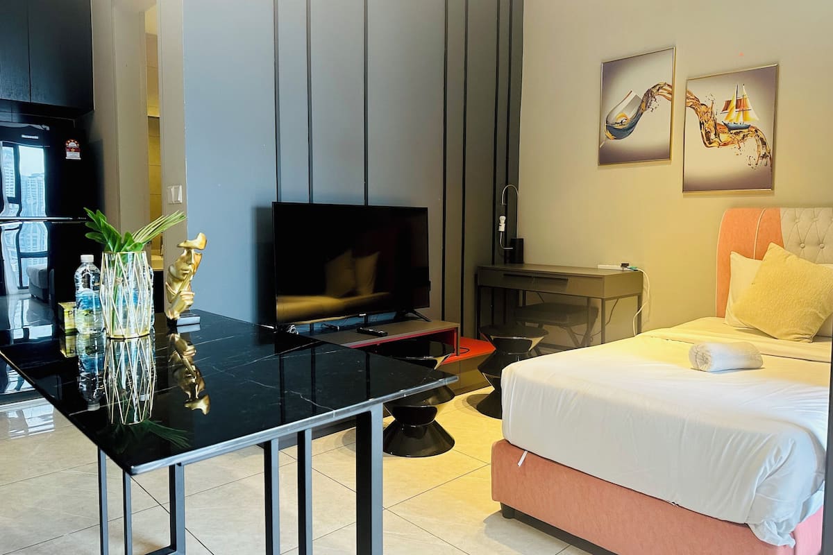 A modern hotel room with a flat-screen TV, a bed with a pink headboard, a desk with a chair, and a small table with a vase and a statue.