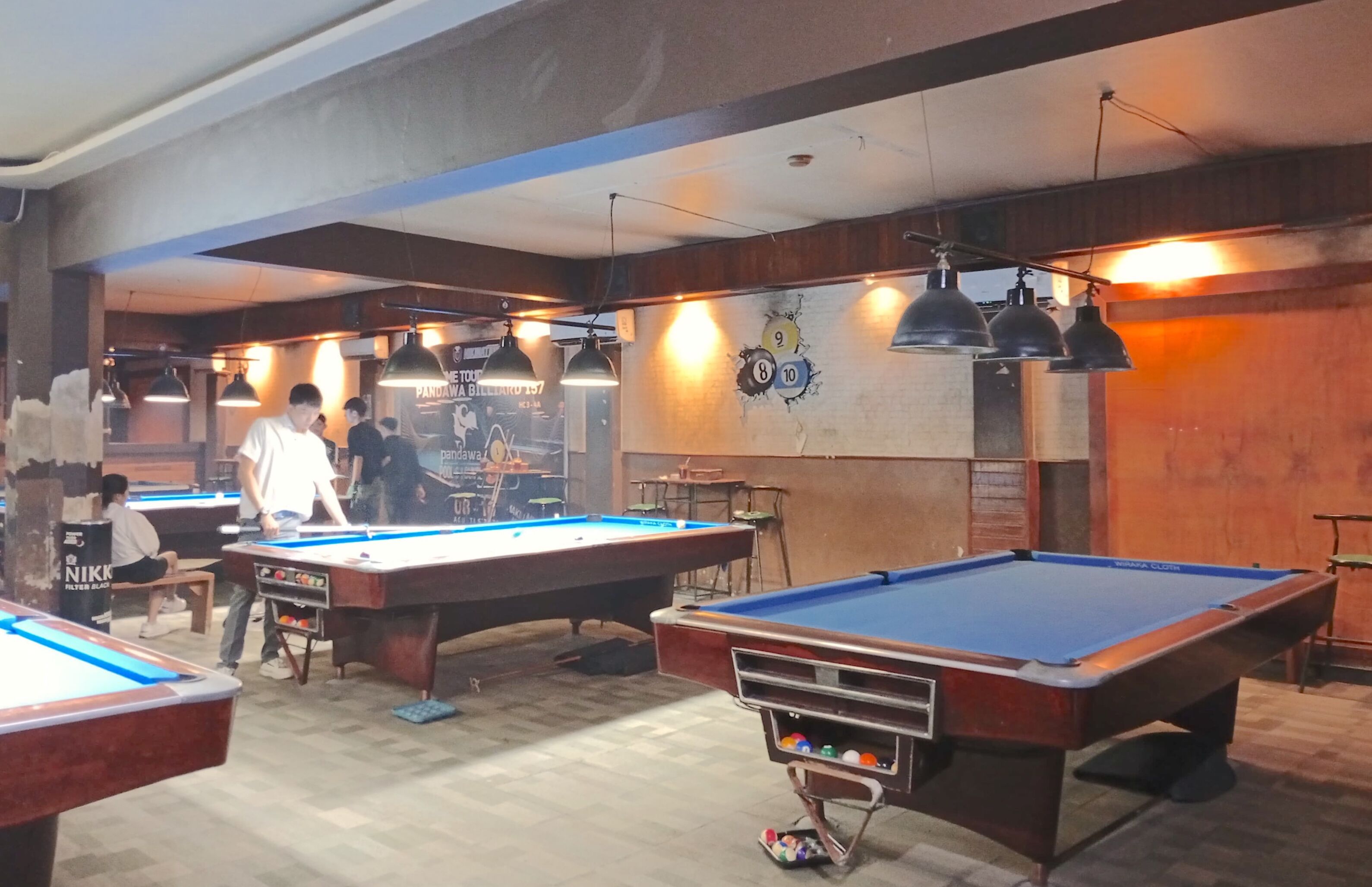 A billiards room with multiple pool tables, overhead lighting, and a mural on the wall.