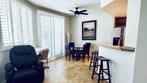 A spacious living area with a black recliner, a dining table with chairs, a kitchen area with a counter and stools, and a ceiling fan.