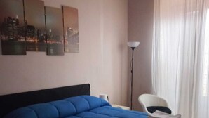 Une chambre d’hôtel avec un grand lit, une œuvre d’art représentant un paysage urbain accrochée au mur, un climatiseur, une lampe sur pied et un fauteuil.