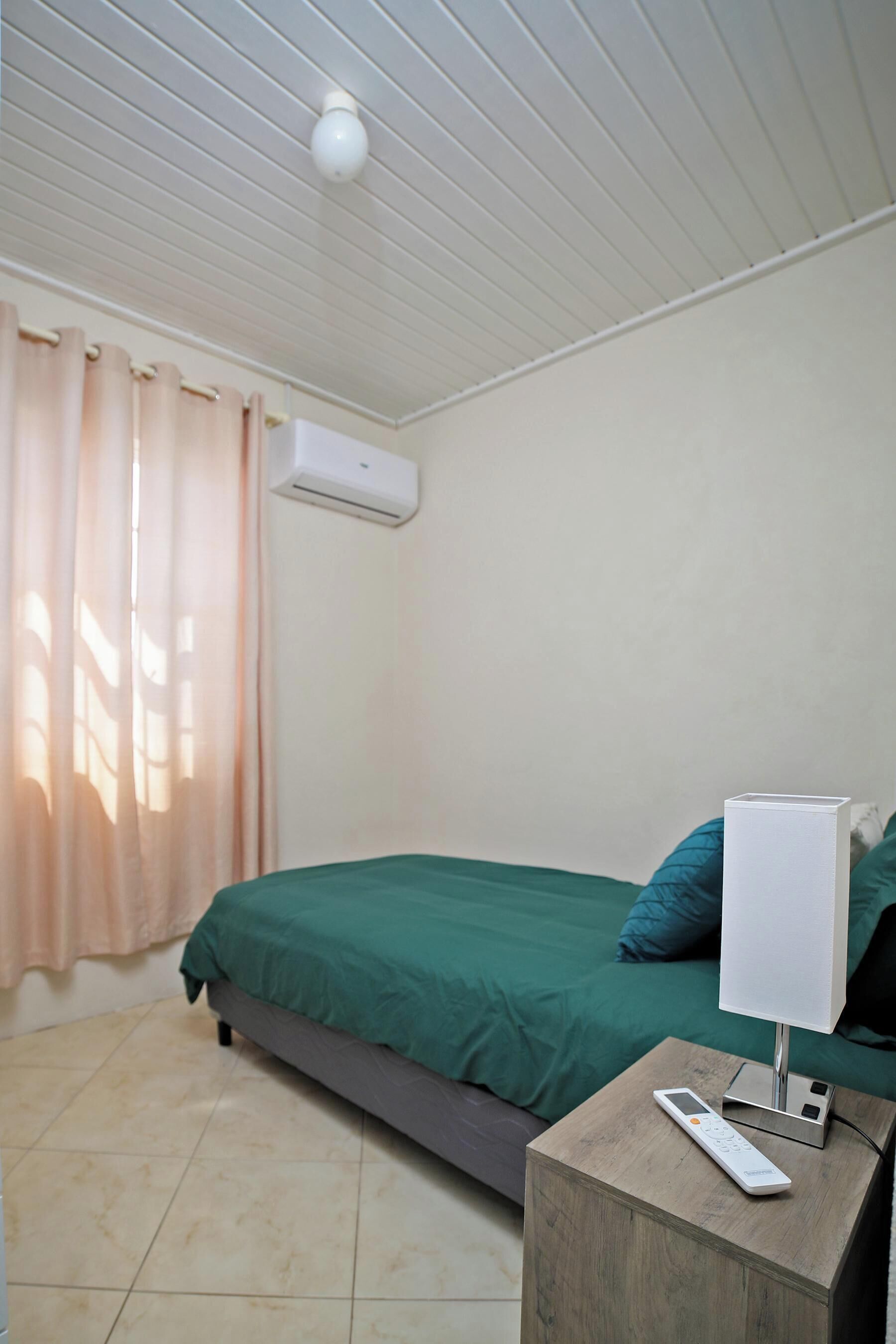 Een slaapkamer met een bed, een nachtkastje met een afstandsbediening, een airconditioner en een raam met gordijnen.