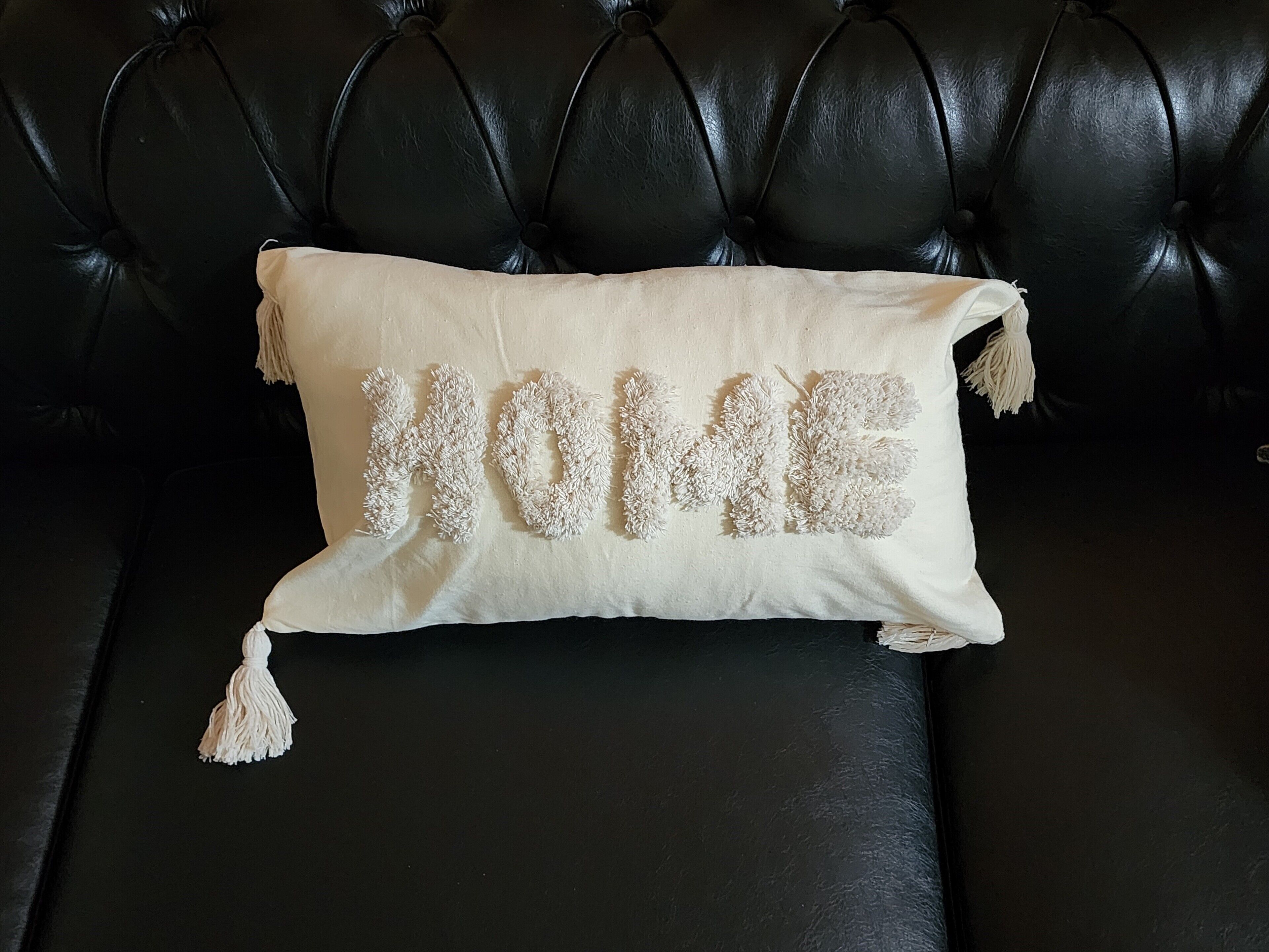 Een zwarte leren bank met een beige kussen waarop het woord "HOME" staat in een getextureerd, pluizig ontwerp.