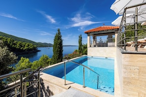 Une piscine avec vue sur un lac et des collines, un espace salon couvert et une terrasse en pierre.