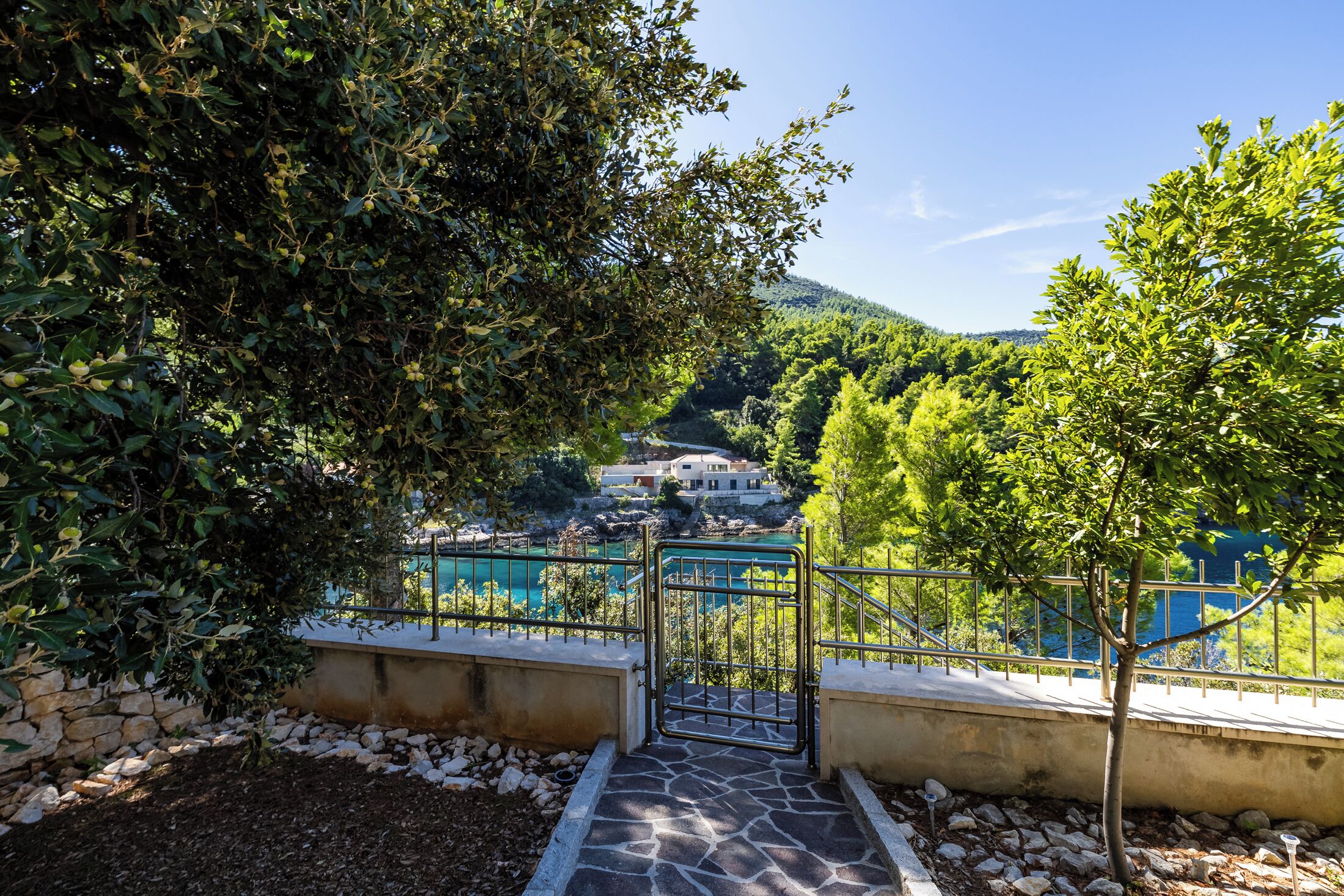 A stone staircase leading to a gate overlooking a coastal area with lush greenery and a view of a building in the distance.