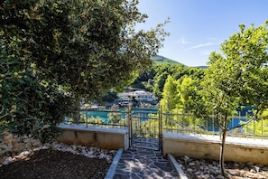 Un escalier en pierre mène à une porte qui donne sur une zone côtière, caractérisée par une végétation luxuriante, et d’où l’on aperçoit un bâtiment au loin.