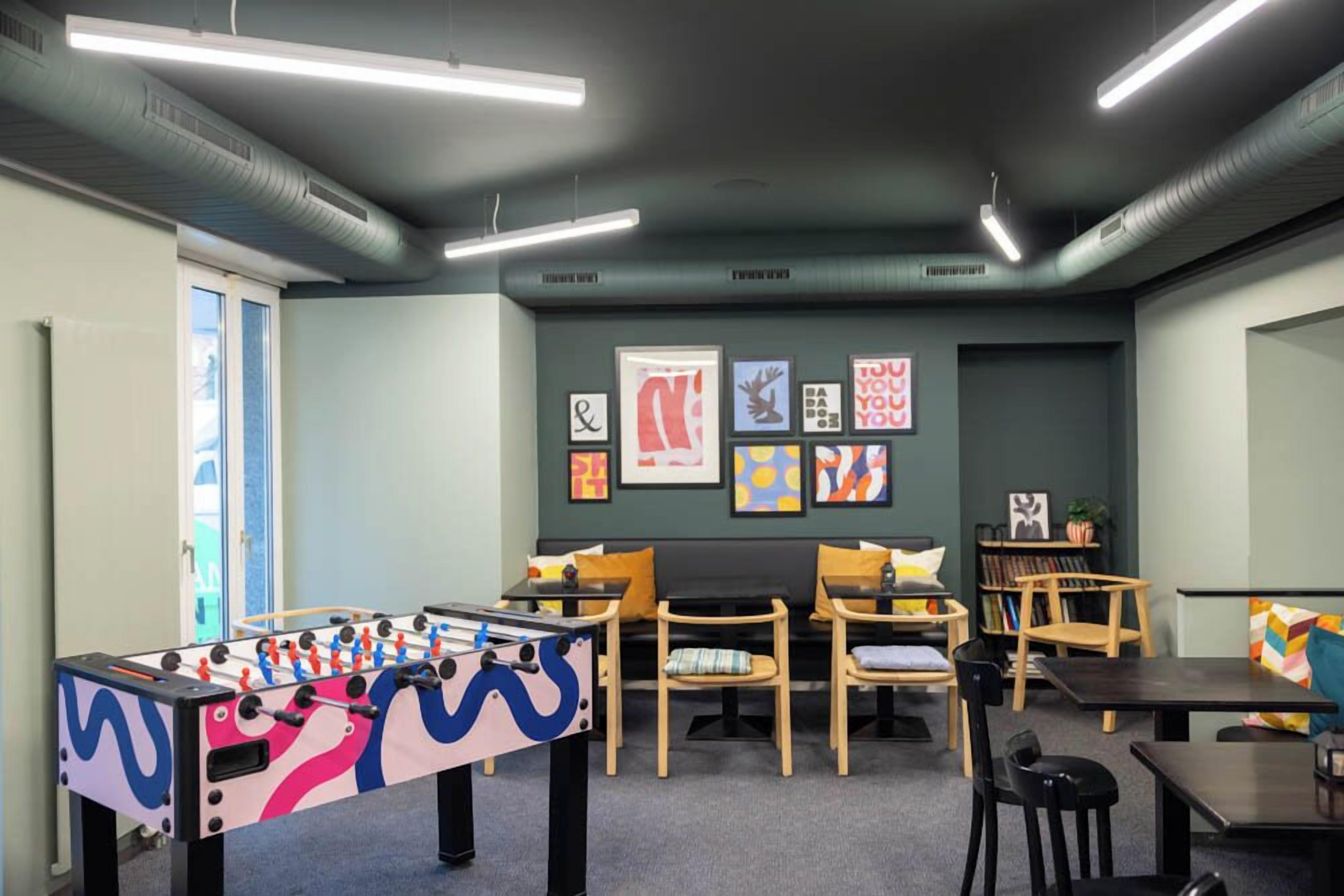 A room with a foosball table, several wooden chairs, tables, and artwork on the walls.