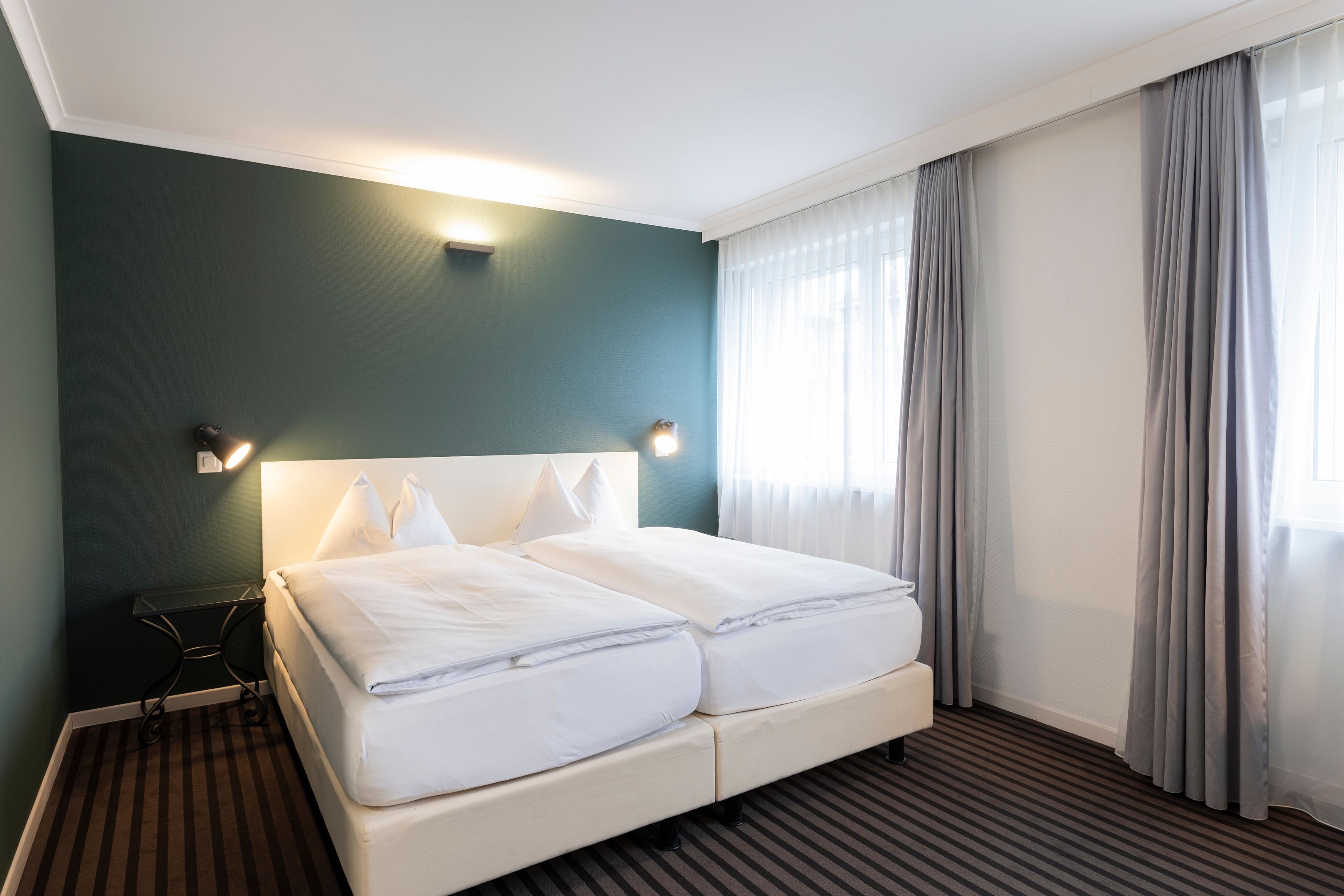 Una habitación de hotel con una cama grande, dos mesitas de noche y un ventanal con cortinas.