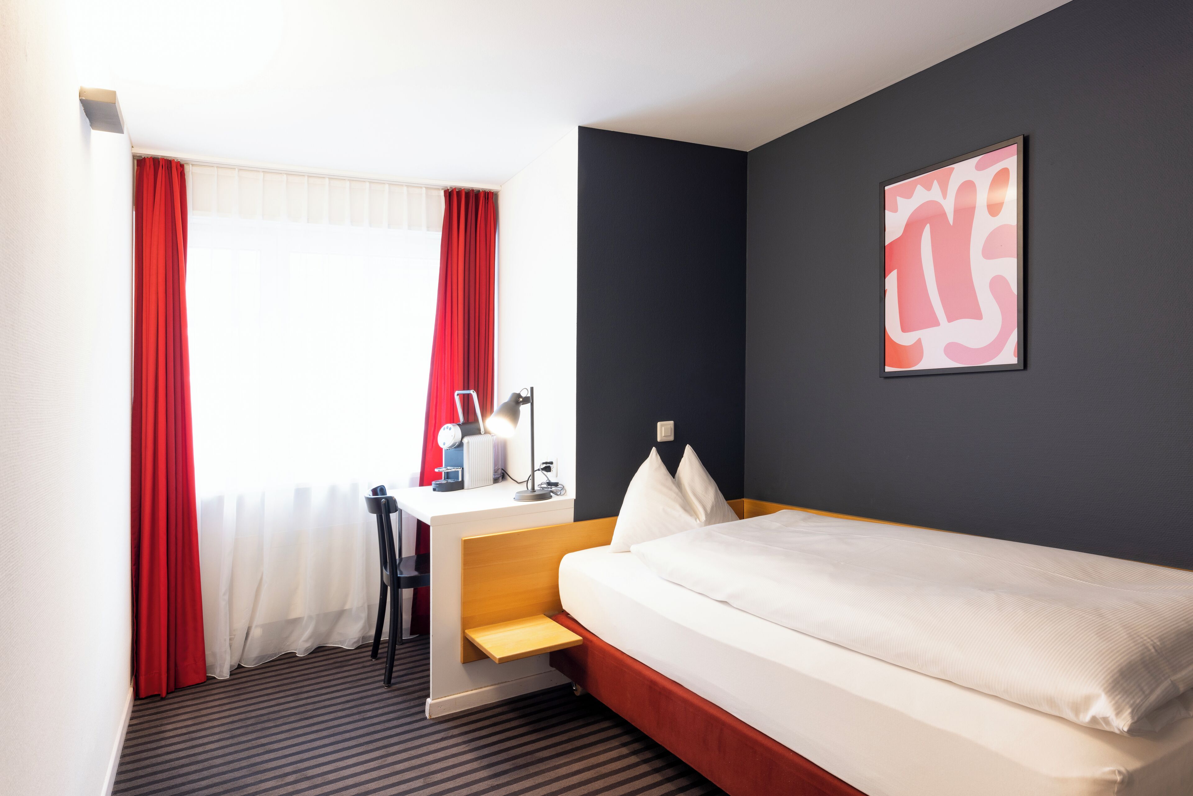 Une chambre d’hôtel avec un lit, un bureau, une chaise et une fenêtre avec des rideaux rouges.